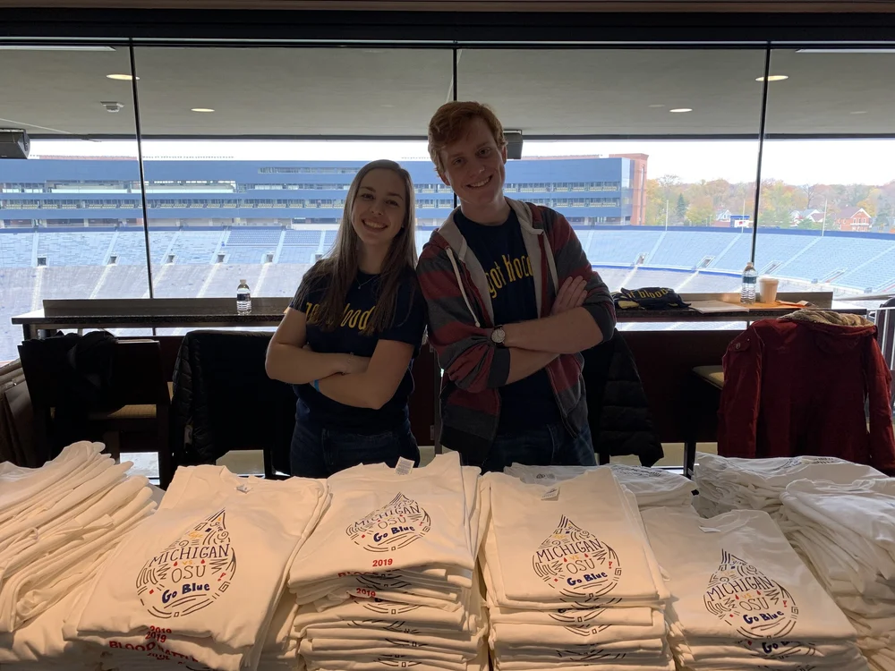 Two BDU volunteers behind stacks of shirts overlooking Michigan Stadium